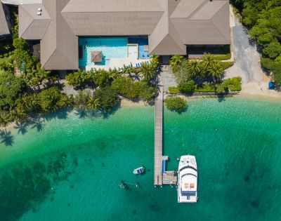 Aerial view of a tropical resort with a large building, pool, and lush greenery. A wooden dock extends to a white yacht on clear turquoise water.