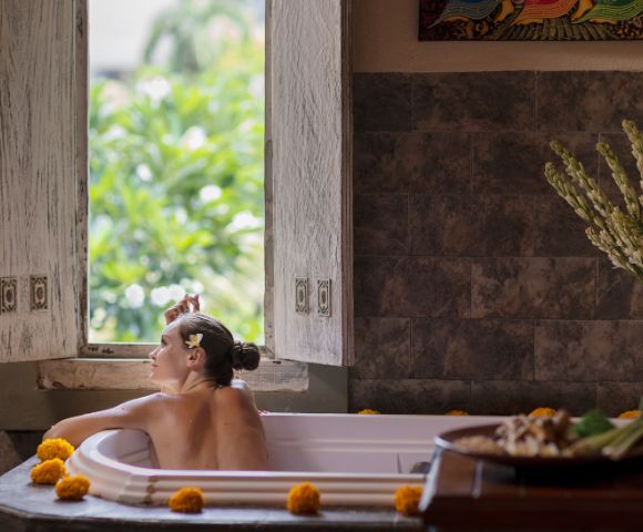Woman with flower in her hair enjoys a bath, looking out open window with decorative shutters, marigolds around the tub's edge, and decoratively arranged plants and flowers on nearby table.
