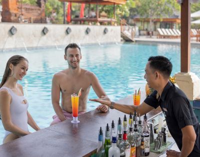 A couple seated in a swimming pool at a swim-up bar, smiling and interacting with the bartender, who is handing them cocktails.