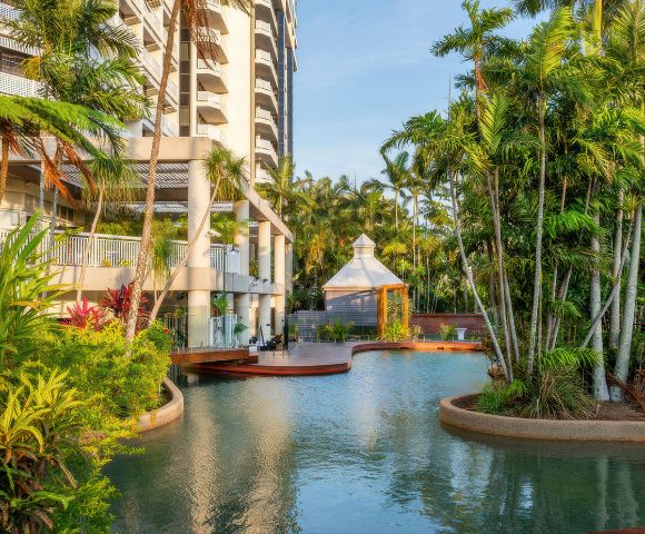Exterior of a resort featuring a lagoon-style swimming pool surrounded by lush tropical landscaping, palm trees, and a gazebo, with a high-rise building in the background.