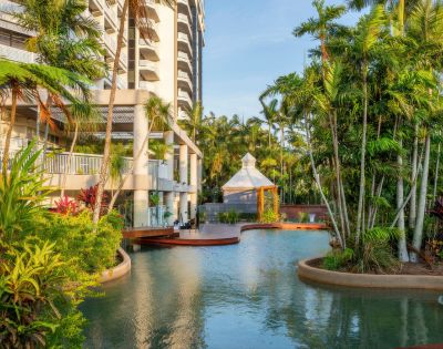 Exterior of a resort featuring a lagoon-style swimming pool surrounded by lush tropical landscaping, palm trees, and a gazebo, with a high-rise building in the background.