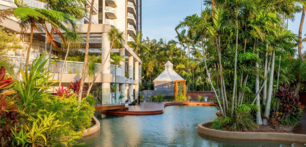 Exterior of a resort featuring a lagoon-style swimming pool surrounded by lush tropical landscaping, palm trees, and a gazebo, with a high-rise building in the background.
