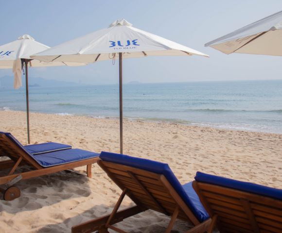 Sunlit beach scene with empty lounge chairs under white umbrellas beside gentle ocean waves. Soft blue sky and distant mountains in the background. Calm ambiance.