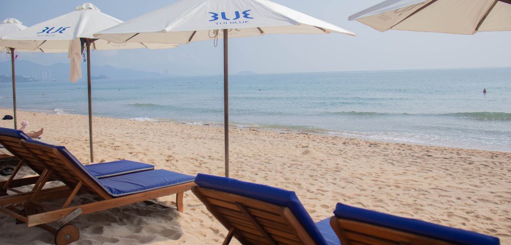 Sunlit beach scene with empty lounge chairs under white umbrellas beside gentle ocean waves. Soft blue sky and distant mountains in the background. Calm ambiance.