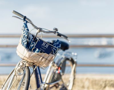 Blue bicycle with a front basket on a sunny seaside path. The basket contains a patterned cloth. The blurred ocean view adds a serene, relaxed vibe.
