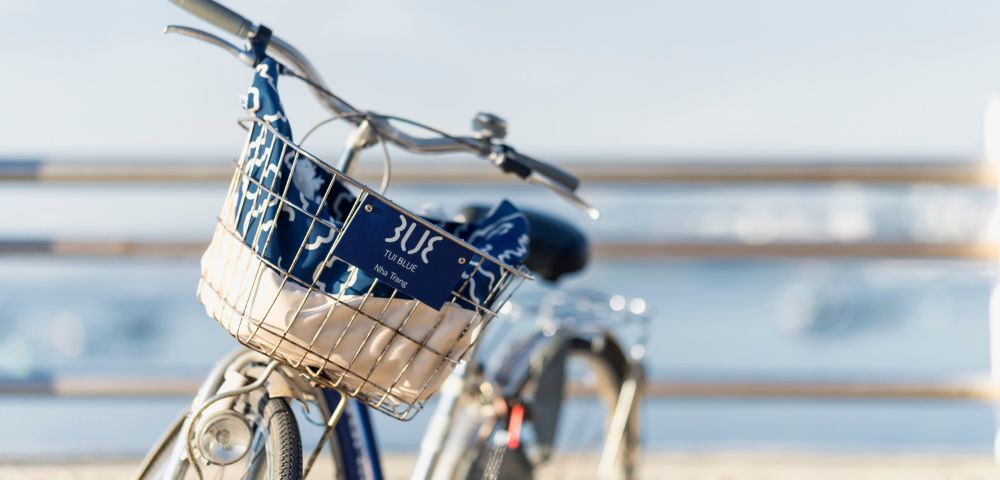 Blue bicycle with a front basket on a sunny seaside path. The basket contains a patterned cloth. The blurred ocean view adds a serene, relaxed vibe.