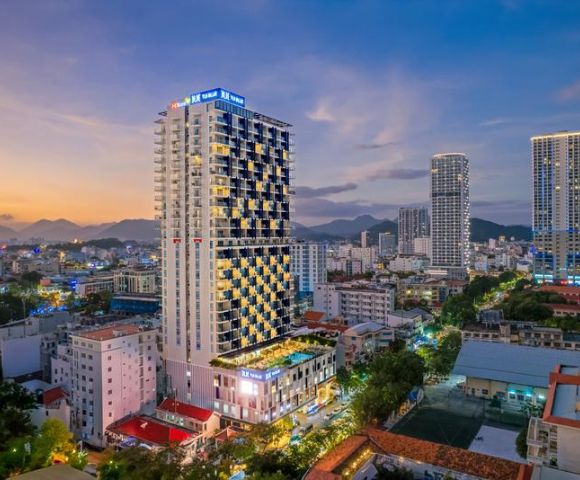 Aerial view of a vibrant cityscape at dusk, featuring a tall, illuminated hotel with a pool. Surrounding buildings are lit against a backdrop of mountains.