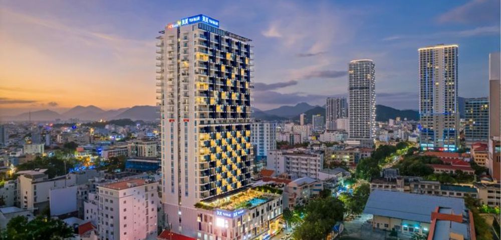 Aerial view of a vibrant cityscape at dusk, featuring a tall, illuminated hotel with a pool. Surrounding buildings are lit against a backdrop of mountains.