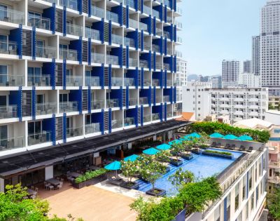 Modern hotel with a blue and white facade, featuring balconies. A rooftop pool with turquoise umbrellas below overlooks a cityscape with tall buildings.