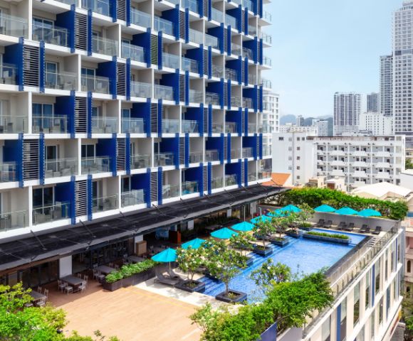 Modern hotel with a blue and white facade, featuring balconies. A rooftop pool with turquoise umbrellas below overlooks a cityscape with tall buildings.