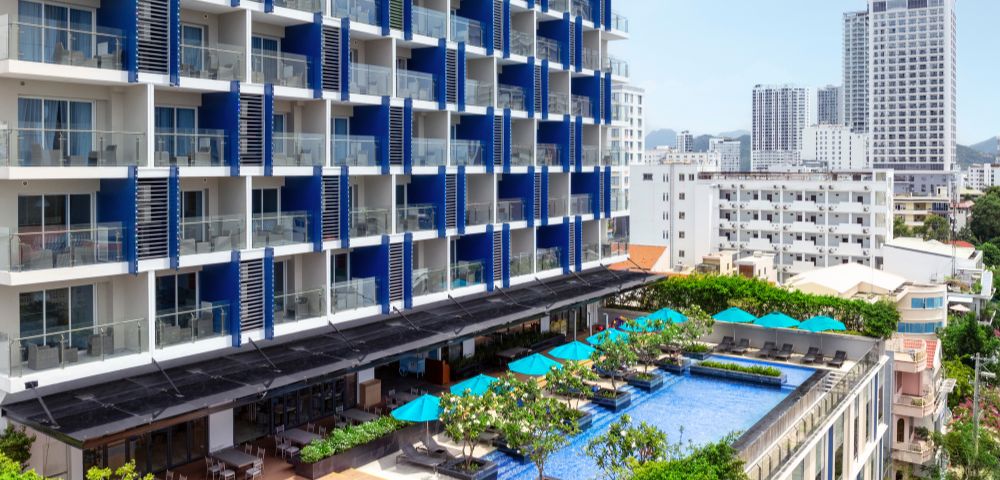 Modern hotel with a blue and white facade, featuring balconies. A rooftop pool with turquoise umbrellas below overlooks a cityscape with tall buildings.