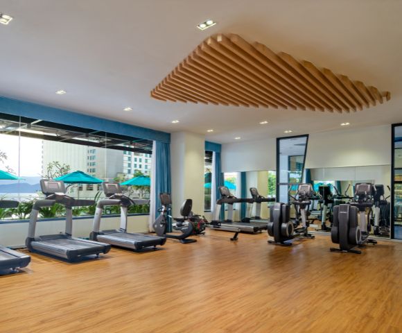 Spacious gym with wooden floors, six treadmills, and exercise bikes by large windows. Bright, modern design with a calm, inviting atmosphere.