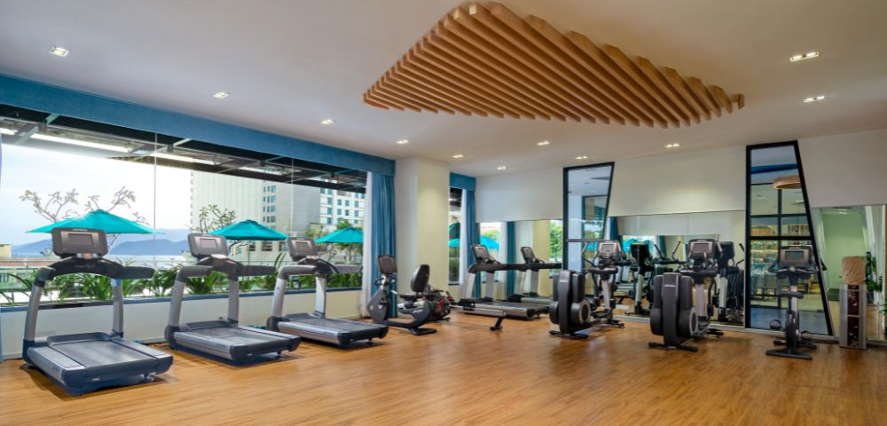 Spacious gym with wooden floors, six treadmills, and exercise bikes by large windows. Bright, modern design with a calm, inviting atmosphere.