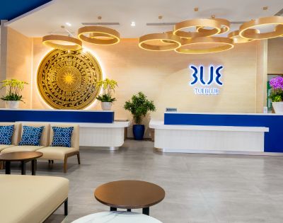 Modern hotel lobby with a blue and white reception desk, decorative wooden ceiling lights, a large ornate circular wall art, and plush seating.