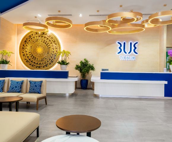 Modern hotel lobby with a blue and white reception desk, decorative wooden ceiling lights, a large ornate circular wall art, and plush seating.