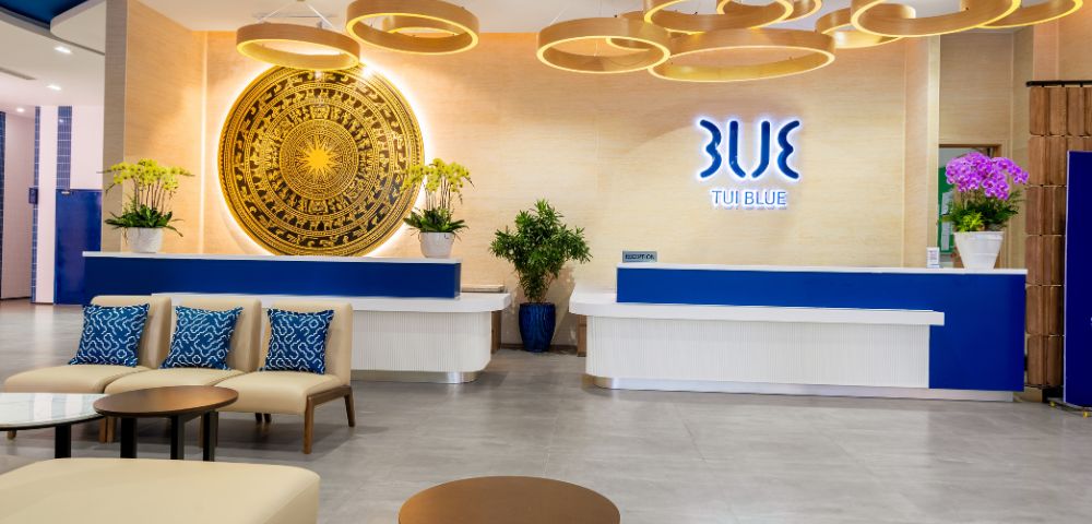 Modern hotel lobby with a blue and white reception desk, decorative wooden ceiling lights, a large ornate circular wall art, and plush seating.