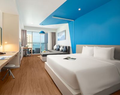 Modern hotel room with a large bed, vibrant blue accent wall, and warm wood flooring. A desk with a lamp is to the side, and a cozy sofa faces a window with an ocean view, enhancing a serene atmosphere.