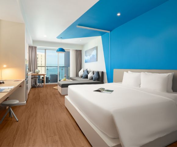 Modern hotel room with a large bed, vibrant blue accent wall, and warm wood flooring. A desk with a lamp is to the side, and a cozy sofa faces a window with an ocean view, enhancing a serene atmosphere.