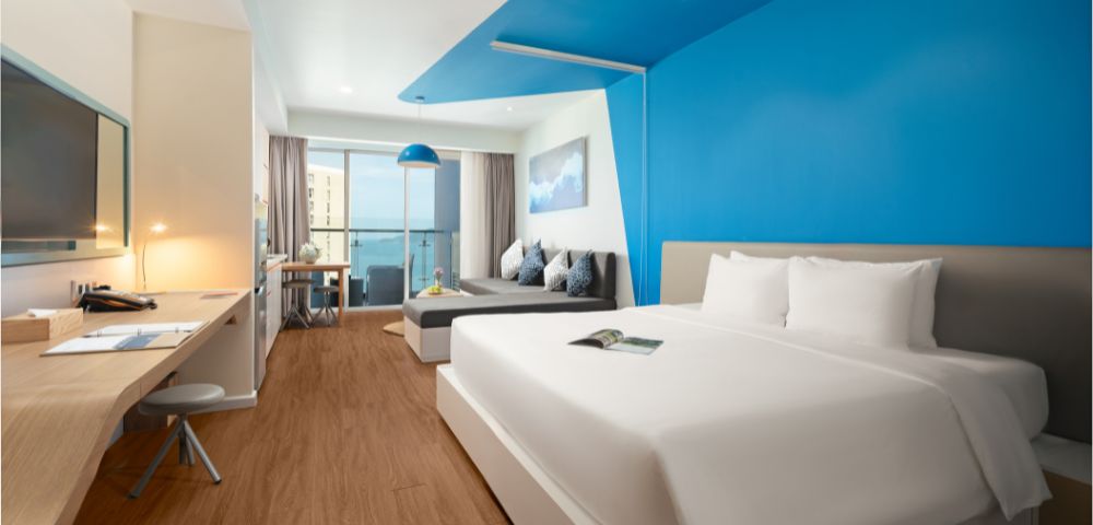 Modern hotel room with a large bed, vibrant blue accent wall, and warm wood flooring. A desk with a lamp is to the side, and a cozy sofa faces a window with an ocean view, enhancing a serene atmosphere.