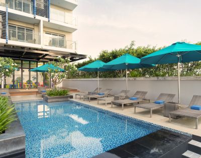 Rooftop pool area with blue mosaic tiles, lounge chairs, and teal umbrellas. Modern building facade in the background; serene and inviting atmosphere.