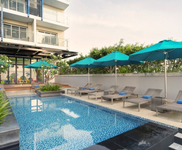 Rooftop pool area with blue mosaic tiles, lounge chairs, and teal umbrellas. Modern building facade in the background; serene and inviting atmosphere.