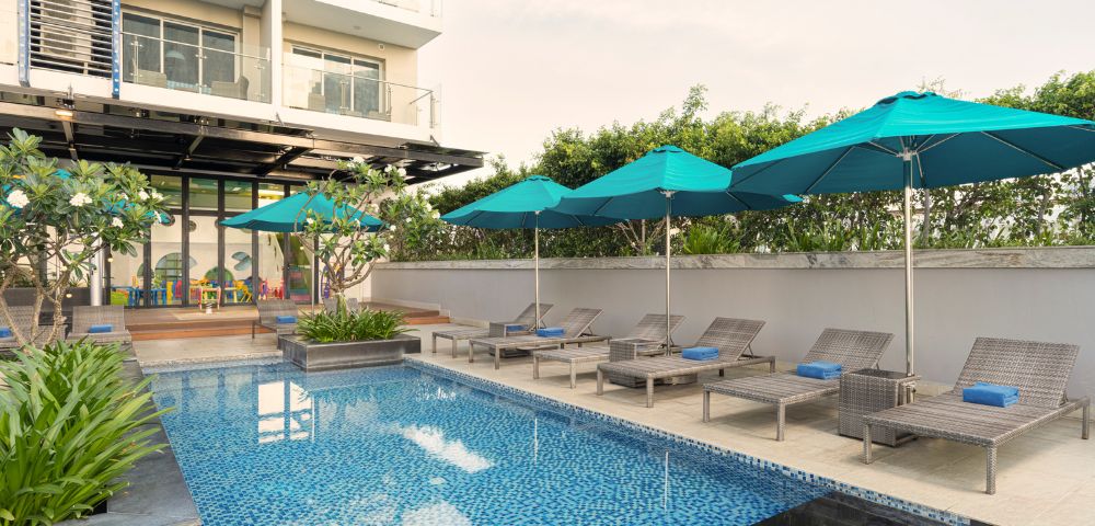 Rooftop pool area with blue mosaic tiles, lounge chairs, and teal umbrellas. Modern building facade in the background; serene and inviting atmosphere.