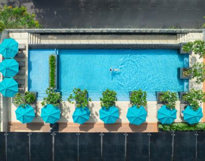 Aerial view of a serene pool with a lone swimmer, surrounded by turquoise umbrellas and lush greenery on a sunny day. The scene is vibrant and tranquil.