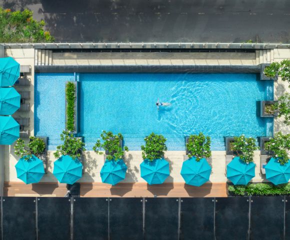 Aerial view of a serene pool with a lone swimmer, surrounded by turquoise umbrellas and lush greenery on a sunny day. The scene is vibrant and tranquil.