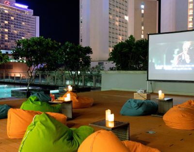 Rooftop cinema at night with colorful bean bags and lit candles, facing a large screen playing a movie. Tall buildings and a pool are in the background.
