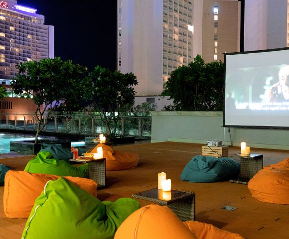 Rooftop cinema at night with colorful bean bags and lit candles, facing a large screen playing a movie. Tall buildings and a pool are in the background.