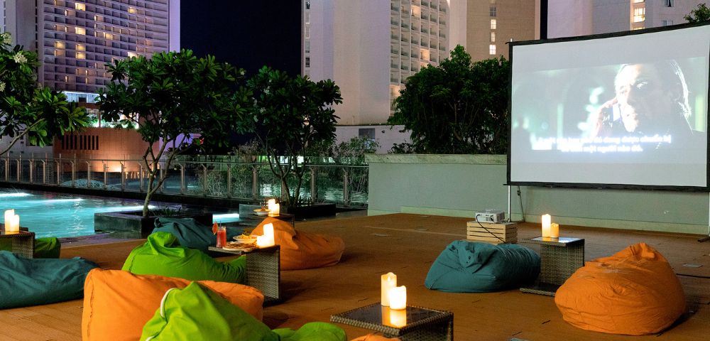 Rooftop cinema at night with colorful bean bags and lit candles, facing a large screen playing a movie. Tall buildings and a pool are in the background.