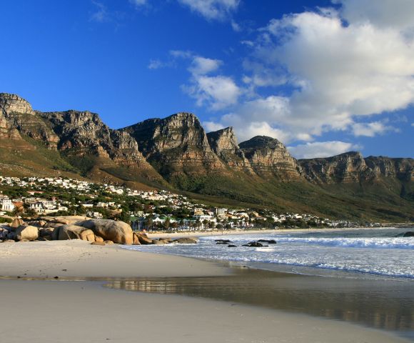 Scenic coastal view of a sandy beach with gentle waves, backed by boulders and hillside houses. Majestic mountains under a bright blue sky with clouds.