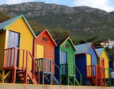 Colorful beach huts in yellow, red, blue, and green line the shore, with a mountainous backdrop. A person in casual attire stands nearby, adding a sense of scale.