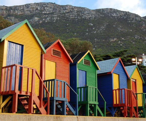 Colorful beach huts in yellow, red, blue, and green line the shore, with a mountainous backdrop. A person in casual attire stands nearby, adding a sense of scale.