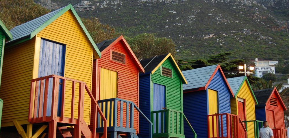 Colorful beach huts in yellow, red, blue, and green line the shore, with a mountainous backdrop. A person in casual attire stands nearby, adding a sense of scale.