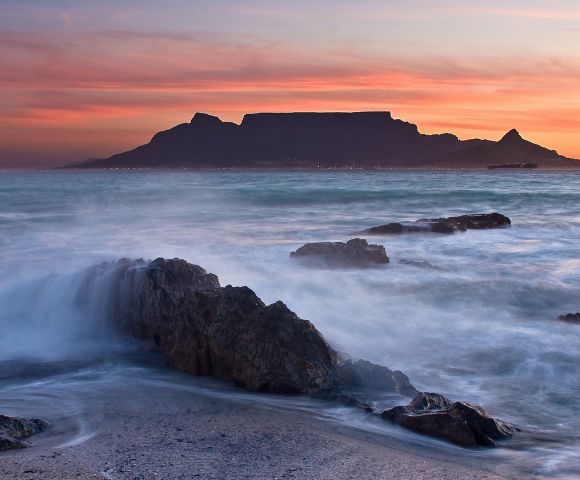 Sunset over ocean with blurred waves crashing against rocks in foreground. Dark silhouette of mountainous horizon under a pink and orange sky. Calming atmosphere.