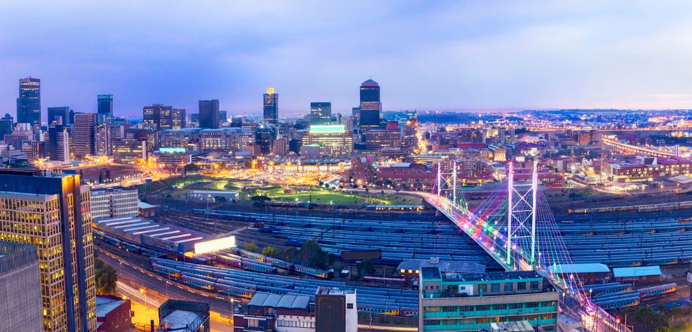 Panoramic view of a vibrant cityscape at dusk, showcasing illuminated skyscrapers and a colorful bridge over a busy railway, evoking a lively urban ambience.