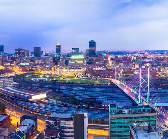 Panoramic view of a vibrant cityscape at dusk, showcasing illuminated skyscrapers and a colorful bridge over a busy railway, evoking a lively urban ambience.