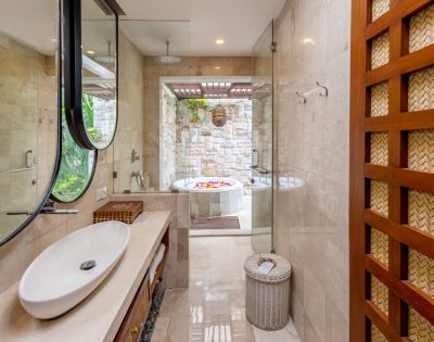 Luxurious spa-like bathroom with a white oval sink atop a stone countertop, oval mirrors with black frames, a glass-enclosed shower, and a deep soaking tub filled with flower petals visible through an opening to an outdoor space. A decorative woven wood paneling is on the right side of the frame.