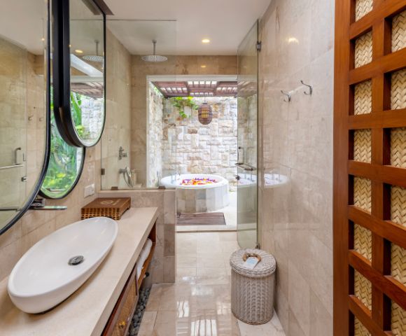 Luxurious spa-like bathroom with a white oval sink atop a stone countertop, oval mirrors with black frames, a glass-enclosed shower, and a deep soaking tub filled with flower petals visible through an opening to an outdoor space. A decorative woven wood paneling is on the right side of the frame.