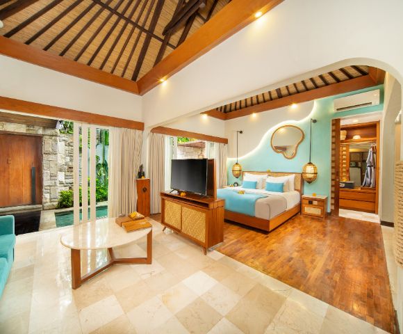 Luxurious open-plan bedroom suite featuring a wooden slatted roof and wood-paneled accents. The room includes a four-poster bed with a turquoise throw and pillows, a wall-mounted mirror, hanging lamps, a TV, wooden furniture, and a turquoise sofa. Large glass doors reveal an outdoor pool area and private entrance. The suite is finished with tile and wood flooring.