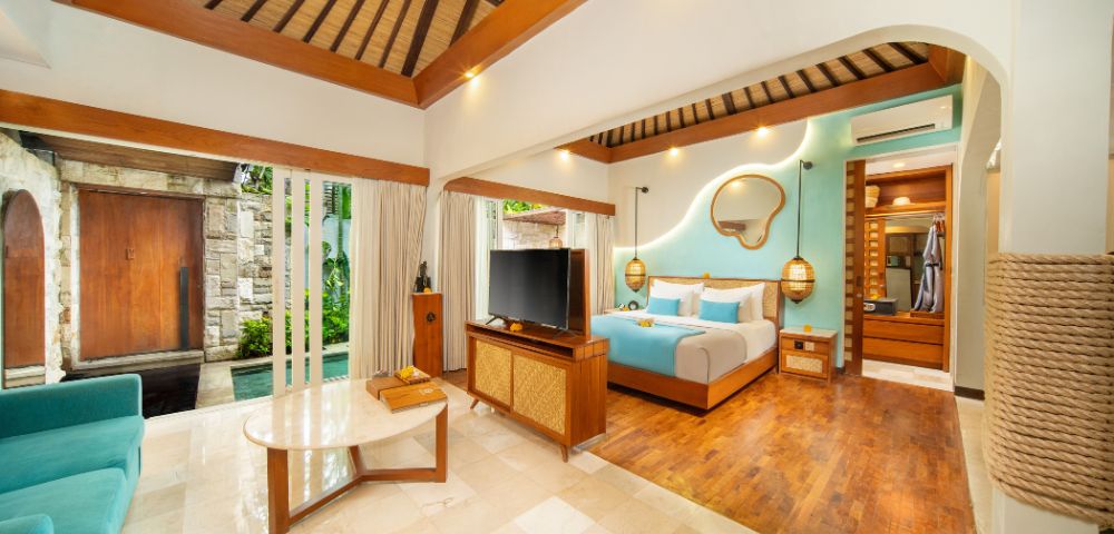 Luxurious open-plan bedroom suite featuring a wooden slatted roof and wood-paneled accents. The room includes a four-poster bed with a turquoise throw and pillows, a wall-mounted mirror, hanging lamps, a TV, wooden furniture, and a turquoise sofa. Large glass doors reveal an outdoor pool area and private entrance. The suite is finished with tile and wood flooring.