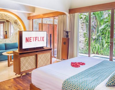 Luxury bedroom interior with a large TV displaying the Netflix logo, a bed with a heart of rose petals, and a view of lush greenery through large windows.