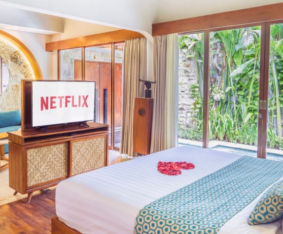 Luxury bedroom interior with a large TV displaying the Netflix logo, a bed with a heart of rose petals, and a view of lush greenery through large windows.
