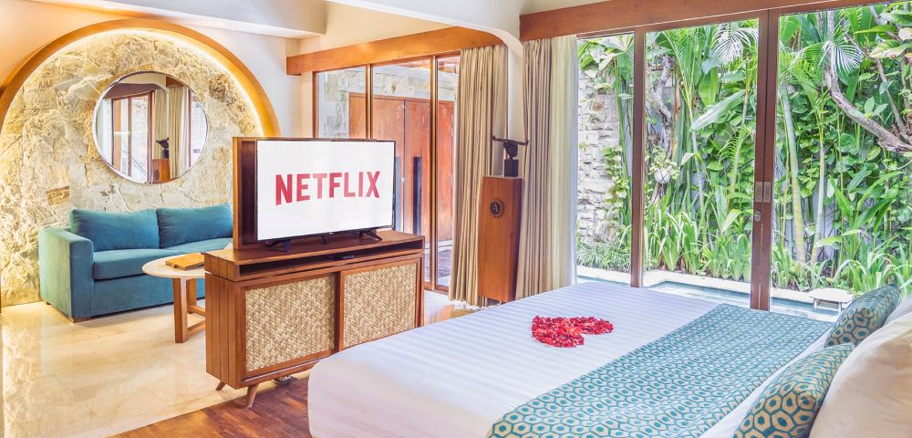Luxury bedroom interior with a large TV displaying the Netflix logo, a bed with a heart of rose petals, and a view of lush greenery through large windows.