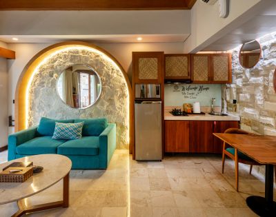 Interior of a modern, open-plan room with a turquoise sofa, a small kitchen, and a dining area; the room is illuminated by warm lighting from a hidden source behind a stone wall and an arched accent over the sofa, with a wooden roof overhead.