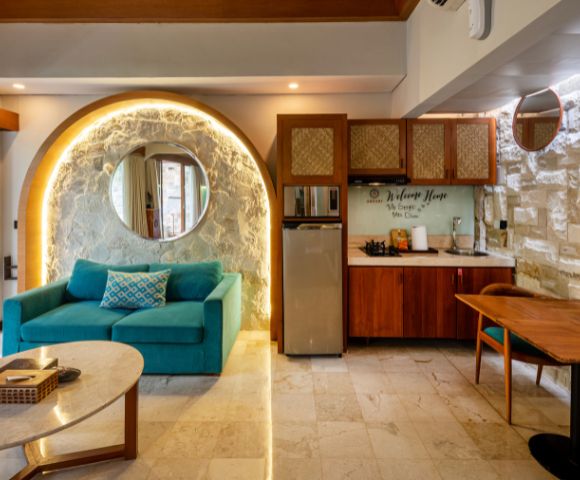 Interior of a modern, open-plan room with a turquoise sofa, a small kitchen, and a dining area; the room is illuminated by warm lighting from a hidden source behind a stone wall and an arched accent over the sofa, with a wooden roof overhead.
