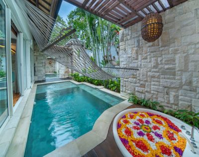 Outdoor spa area with a turquoise plunge pool, a hanging rope hammock, and a stone wall with a white bathtub filled with colorful flower petals. The space is partially covered with a wooden pergola, and features sliding glass doors opening to a bright interior. A woven lantern hangs from the pergola, and lush greenery surrounds the scene.