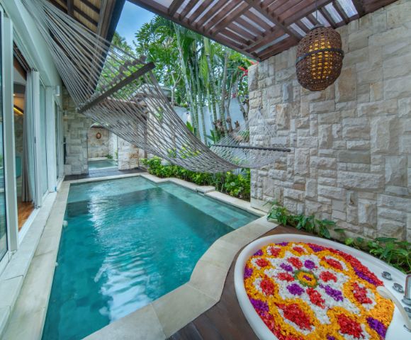 Outdoor spa area with a turquoise plunge pool, a hanging rope hammock, and a stone wall with a white bathtub filled with colorful flower petals. The space is partially covered with a wooden pergola, and features sliding glass doors opening to a bright interior. A woven lantern hangs from the pergola, and lush greenery surrounds the scene.
