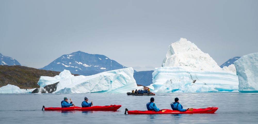 Four kayakers in red paddles explore icy waters, with large icebergs and mountains in the background. A small boat is nearby.